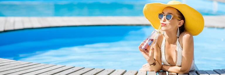 Junge Frau im Badeanzug mit großem gelben Sonnenhut entspannt sich mit einer Flasche frischem Getränk am Pool sitzend im Freien.