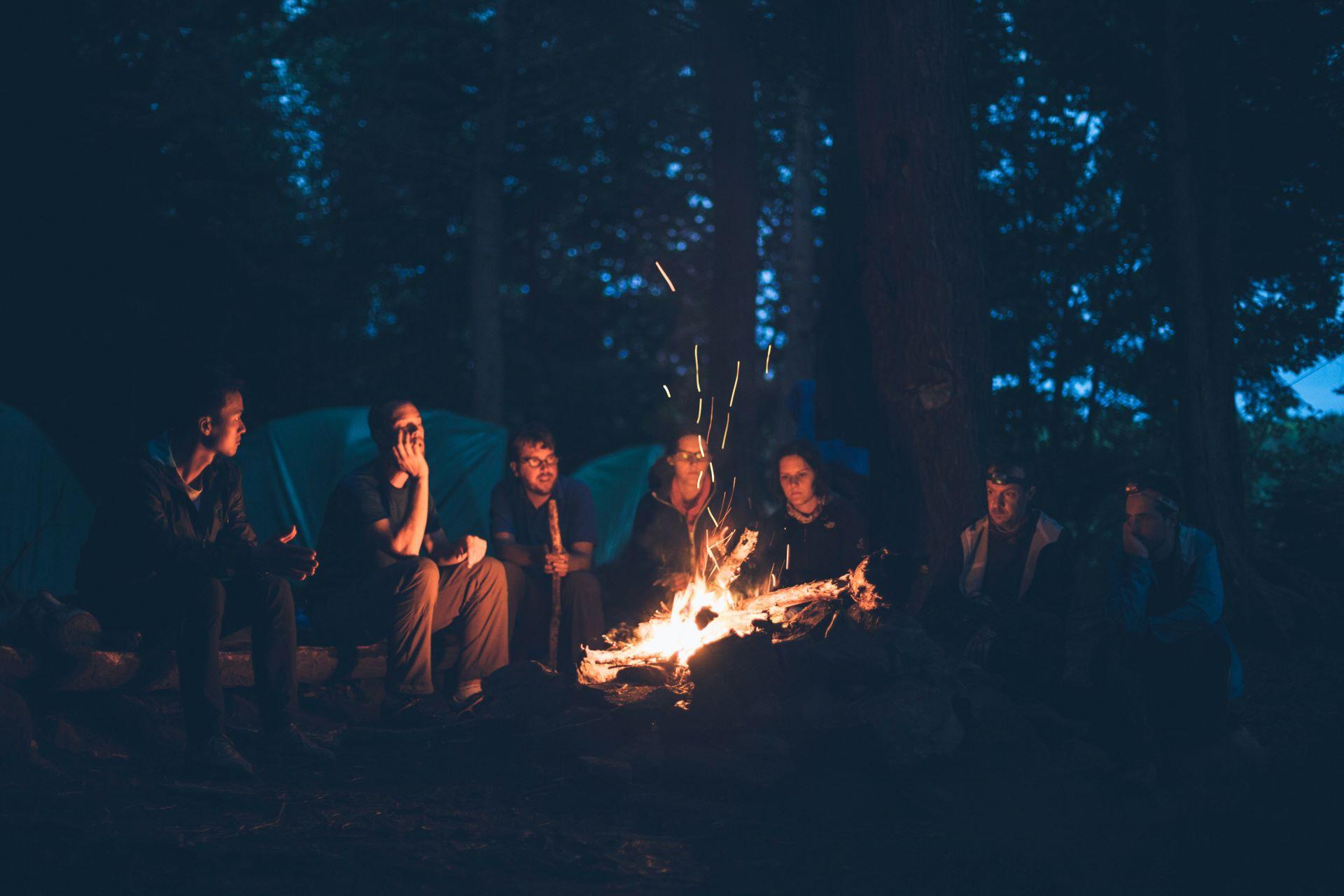 Eine Gruppe von Menschen sitzt um ein Lagerfeuer, umgeben von Zelten – Camping-Atmosphäre bei Nacht.
