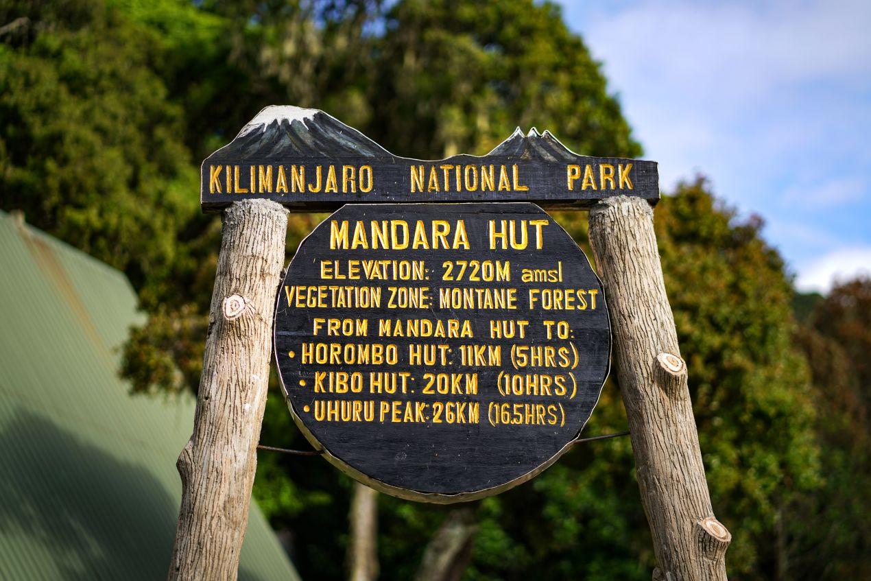 Ein hölzernes Schild der Mandara Hut im Kilimanjaro-Nationalpark, das Wegstrecken und Kilimanjaro Besteigung Kosten symbolisiert.